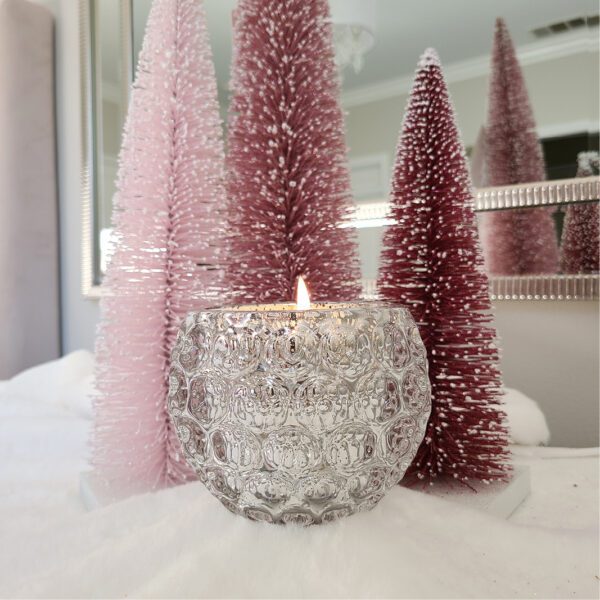 A candle in a glass bowl next to two pink christmas trees.